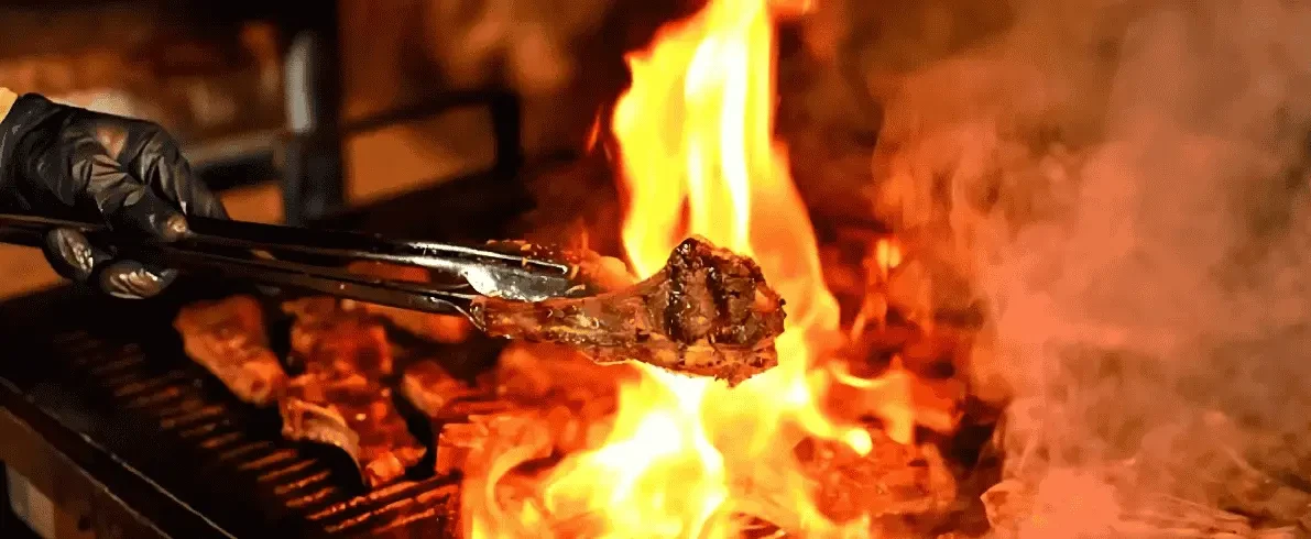At a desert safari BBQ dinner, a person in blue gloves holds tongs over a grill under the night sky.