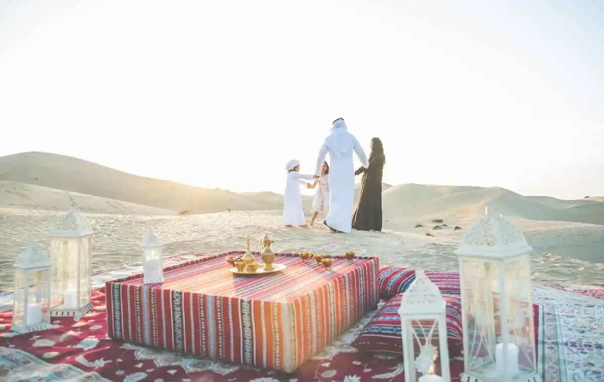 A family enjoys breakfast on a blanket in the desert, surrounded by sand and morning sunlight.