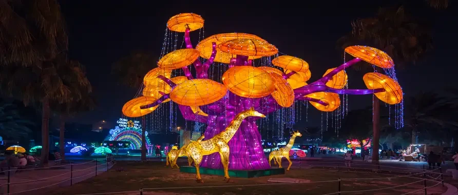 A giraffe stands beside a tree in the Glow Garden, surrounded by vibrant illuminated plants.