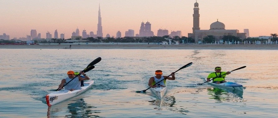 Tourists kayaking on calm waters at sunset with Dubai skyline and mosque in the background.