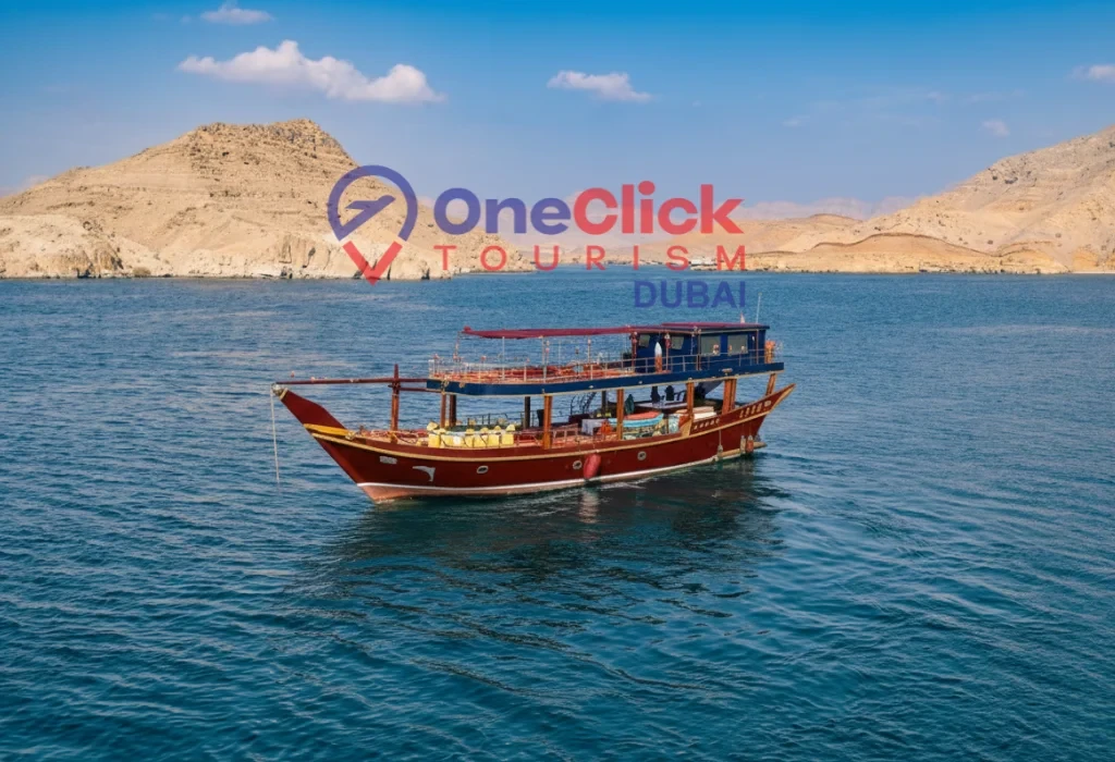A traditional wooden Arabian dhow boat sailing on turquoise sea water with rugged desert mountains and a clear blue sky in the background.