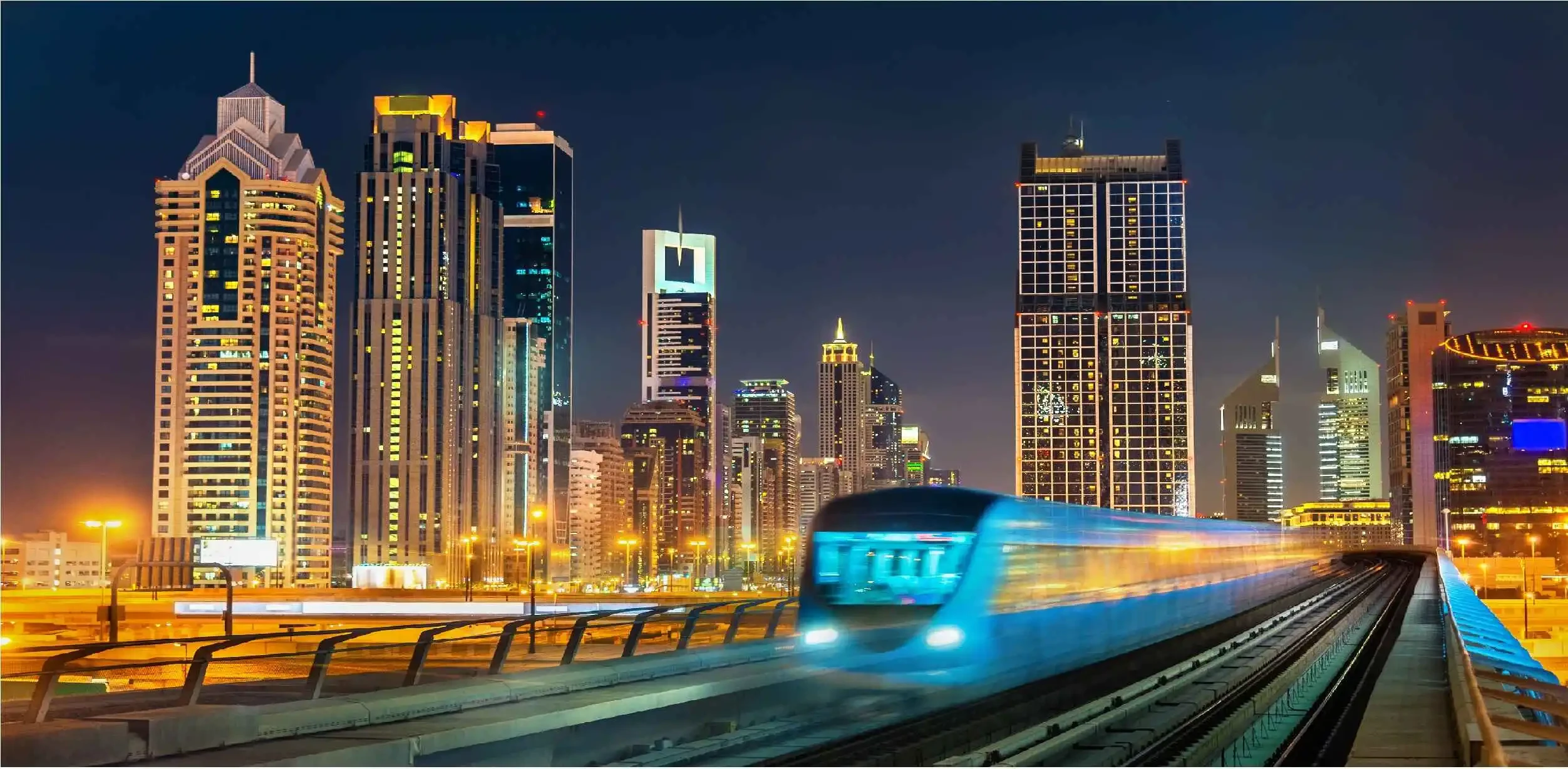 A night view of a train moving through the city on Sheikh Zayed Road, surrounded by glowing buildings and streetlights.