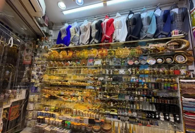 A souvenir shop in a traditional Dubai souk displaying Arabic dresses, decorative items, camel figurines, jewelry, magnets, and cultural handicrafts on crowded shelves.