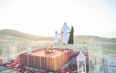 A family enjoys breakfast on a blanket in the desert, surrounded by sand and morning sunlight.