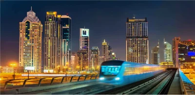 A night view of a train moving through the city on Sheikh Zayed Road, surrounded by glowing buildings and streetlights.
